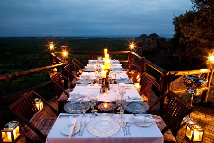 A set dining table on a wooden deck with lit lanterns, overlooking a vast green landscape under a cloudy sky. The table is elegantly arranged with white tablecloths, glassware, and candles, creating a warm and inviting evening ambiance.