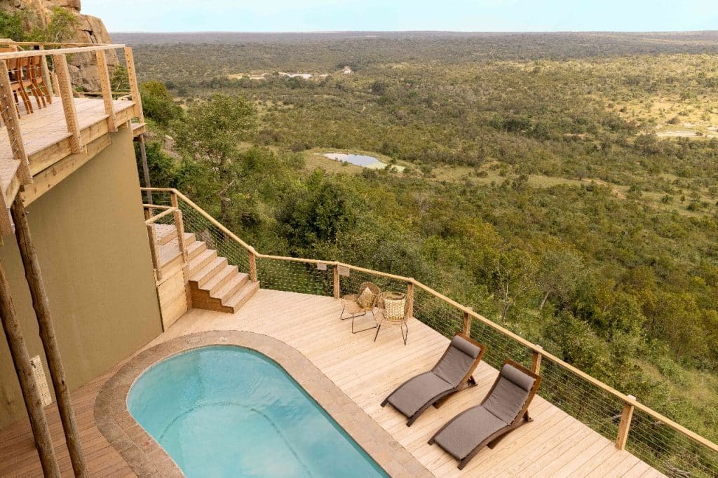 A scenic view from a wooden deck overlooking a vast, green landscape. The deck features a small pool, two lounge chairs, and a table with chairs. The expansive vista includes trees and a distant water feature under a clear sky.