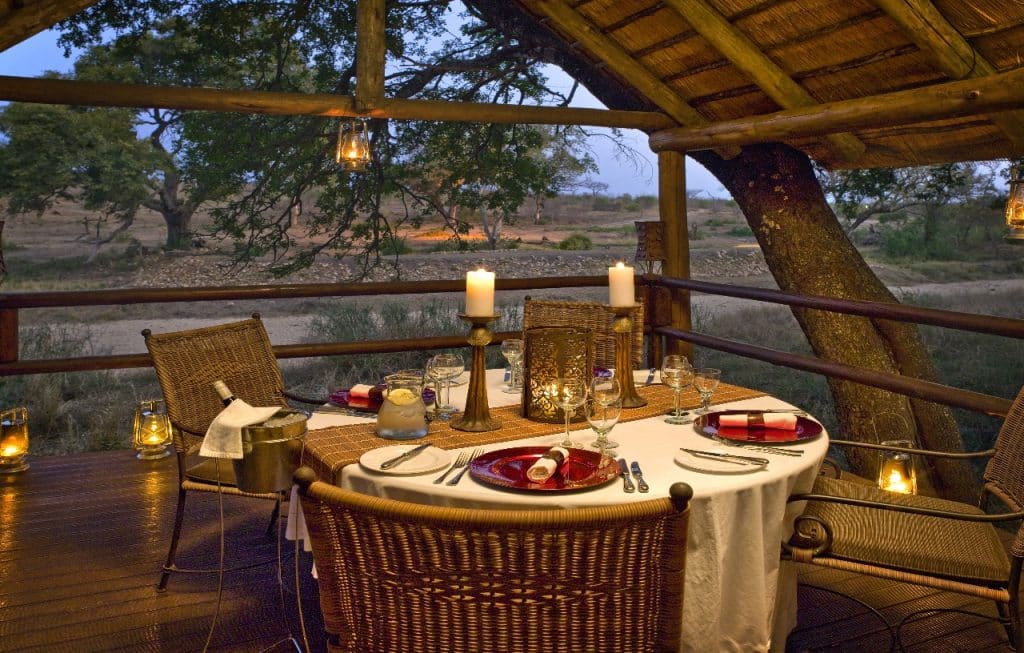 A rustic outdoor dining setup on a wooden deck overlooking a savannah. A round table is elegantly set with red and white napkins, candles, and glassware. Lanterns and a thatched roof add to the ambiance. Trees and open plains are visible in the background.