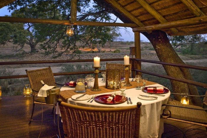 A rustic outdoor dining setup on a wooden deck overlooking a savannah. A round table is elegantly set with red and white napkins, candles, and glassware. Lanterns and a thatched roof add to the ambiance. Trees and open plains are visible in the background.