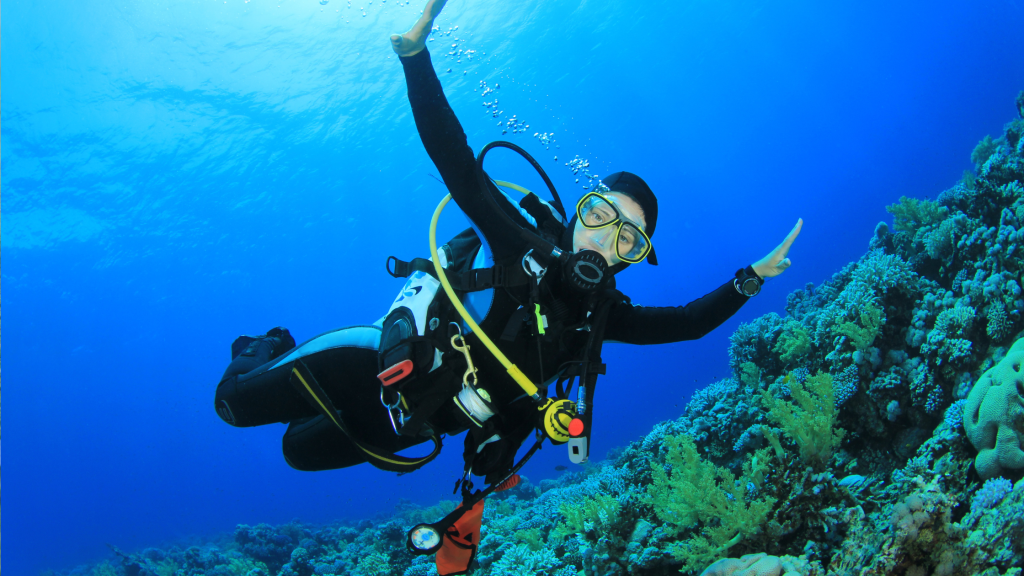 A scuba diver wearing a black wetsuit, yellow mask, and oxygen tank swims underwater near a vibrant coral reef in clear blue water. The diver is surrounded by bubbles and appears to be enjoying the marine environment.