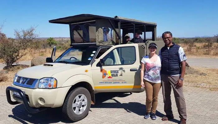 A man and woman stand smiling in front of a safari vehicle with a canopy. The background shows sparse vegetation and a clear sky, suggesting a warm, dry climate.