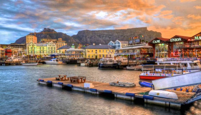 Scenic view of a busy harbor with boats docked along wooden piers, colorful buildings lining the waterfront, and a dramatic sky at sunset. Mountains provide a stunning backdrop, reminiscent of the breathtaking vistas one might find on a 5-day journey from Johannesburg to Cape Town.