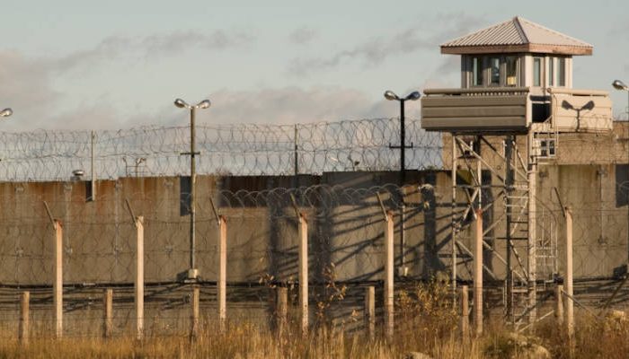 drakenstein A prison yard with a tall perimeter fence topped with barbed wire mirrors the isolation found far from a full day Cape Winelands tour. A guard tower stationed at the corner overlooks the grassy ground, under a partly cloudy sky.