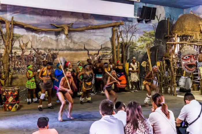 A group of people performs a traditional dance in brightly colored attire, reminiscent of the vibrant displays at Lesedi Cultural Village. The backdrop features an African village scene with huts and a mural. Seated onlookers enjoy the captivating performance in an indoor setting.