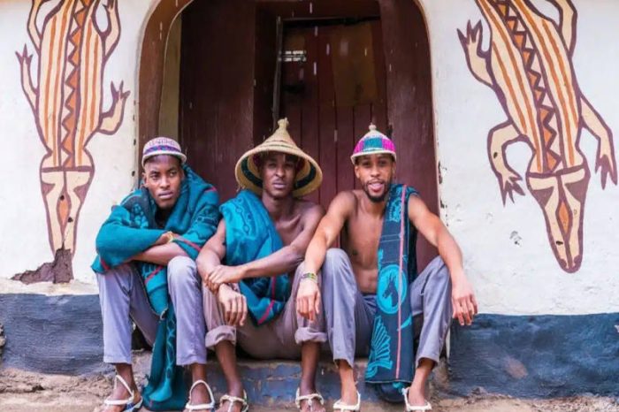 Three men in traditional hats and colorful fabrics sit on a step before a decorated building at Lesedi Cultural Village. The intricate animal designs on the wall frame their relaxed presence, vividly showcasing cultural attire and heritage amidst this vibrant community hub.