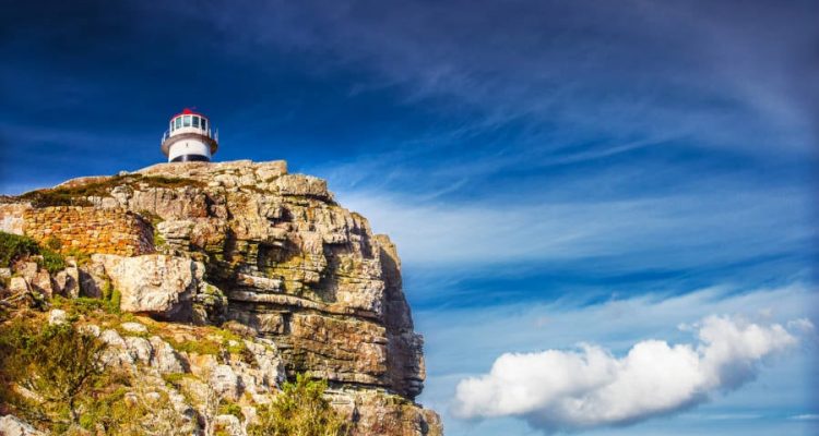 A lighthouse perched atop a rugged cliff under a vivid blue sky with wisps of cloud, much like the scenic views on a Cape Peninsula tour. The landscape is dotted with sparse greenery, conveying solitude and natural beauty.