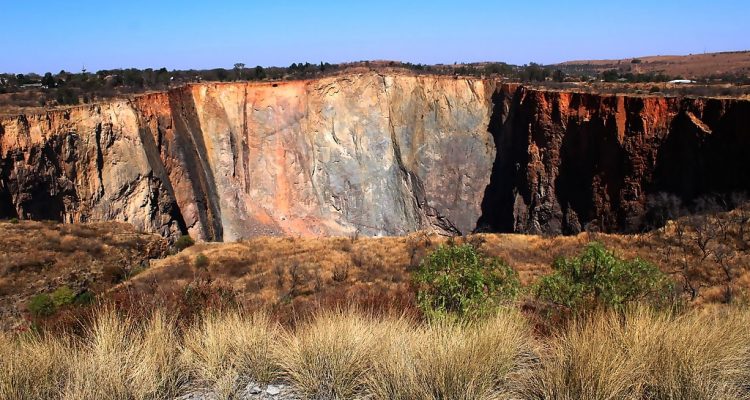 A scenic view unfolds of a large, steep rock face displaying layers of red and gray hues under a clear blue sky. The foreground boasts dry grass, with sparse trees dotting the rugged landscape—a perfect stop on your Pretoria city tour through its arid beauty.