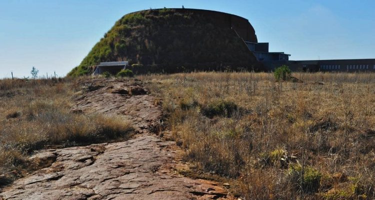 A large, round, grass-covered hill with a stone building at the top stands majestically against a clear blue sky, reminiscent of the landscapes seen on the Cradle of Humankind tour. In the foreground, a rocky pathway winds its way through dry grassland toward the hill.