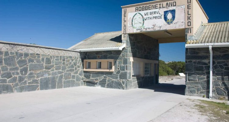 The image captures the entrance to Robben Island, featuring a stone building and an archway labeled "Robben Island Welkom." The sign proudly displays emblems and the slogan "We Serve With Pride" against a clear blue sky, reminiscent of Kruger Tours in Cape Town.